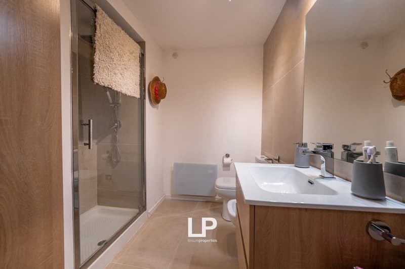 Modern bathroom with walk-in shower, white sink, white tiles, white toilet, white mirror, wooden flooring, and modern fixtures.