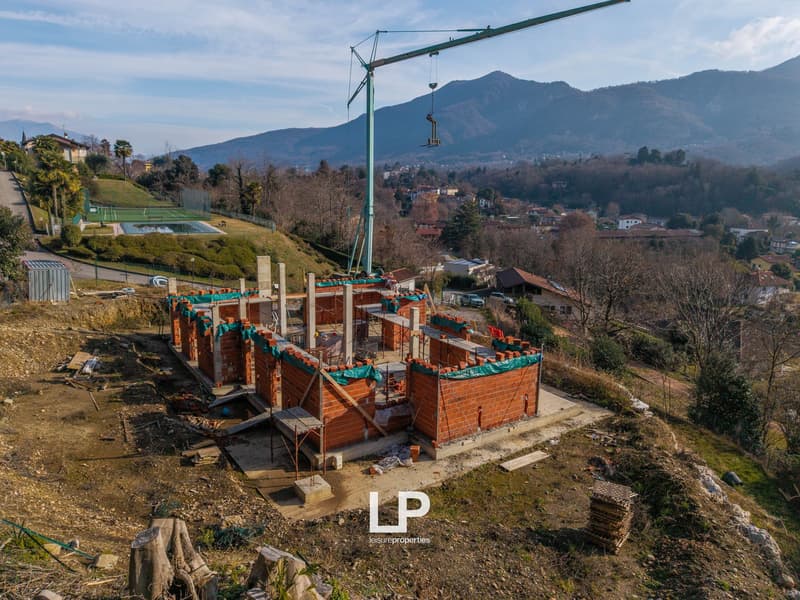 Building under construction, red brick walls, metal scaffolding, crane, mountain backdrop, tennis court, empty field, greenery, houses in the distance