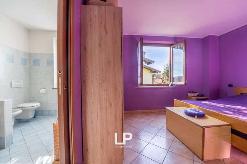 Purple wall, wooden wardrobe, wooden bed with mattress, pillow, wooden bench, bedside table with basket, toilet, sink, toilet paper, light switch