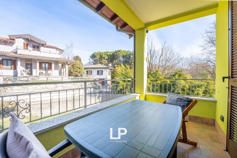 Modern balcony with tile floor, large windows with balcony railings, round table with two chairs, a view of greenery and neighboring houses.