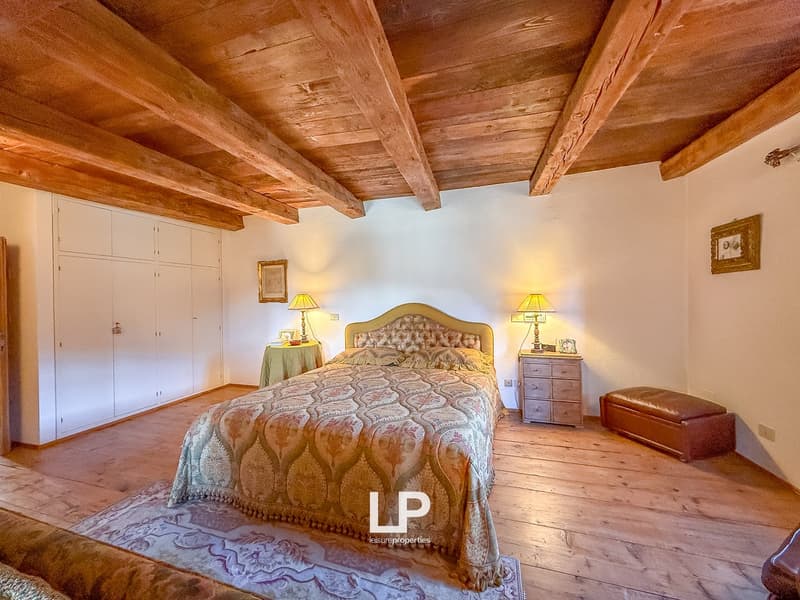 Wooden ceiling, wooden floor, double bed with headboard, two nightstands with lamps, wooden cabinet, large white wardrobe, rug on the floor, bench, picture frame on the wall