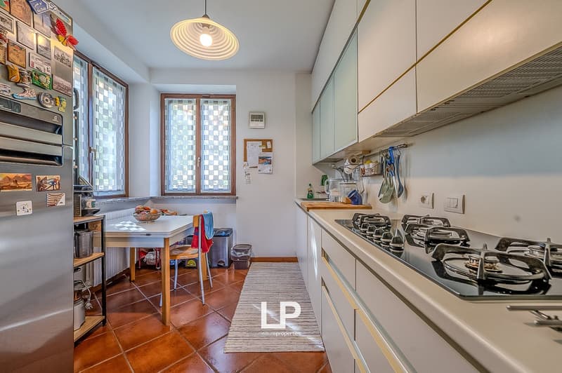 Modern kitchen with gas stove, white cabinets, red tile floor, dining table with chair, refrigerator, lamp, and a trash bin.