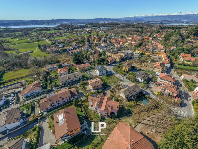 Aerial view of a residential neighborhood with detached houses and apartment buildings, a mix of red roofs, green trees, and small gardens, a serene setting with distant mountains, and a peaceful ambiance.