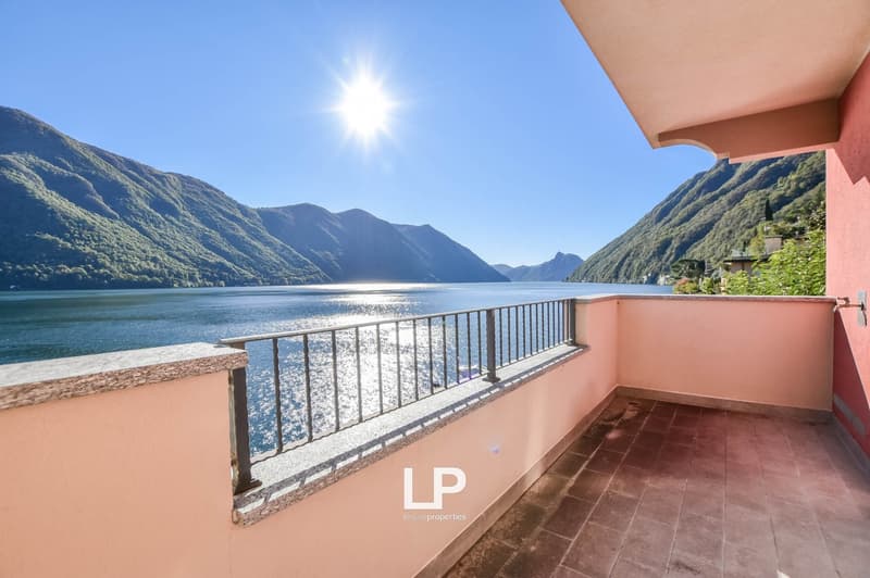 Outdoor balcony with tiled flooring, metal railings, overlooking a serene lake and lush mountains, a glimpse of a house's corner in the distance