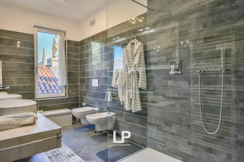 Modern bathroom with gray tiled walls, glass shower enclosure, white bath, toilet, bidet, sink, and a window overlooking a view.