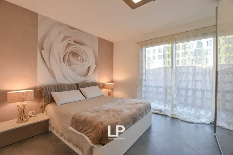 Queen-sized bed, beige comforter, two white pillows, wooden nightstands with lamps, rose wallpaper, sliding doors, white curtains, tile floor, wall-mounted power outlet.