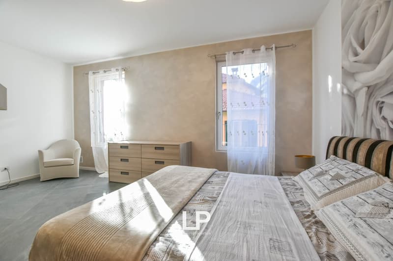 Double bed, two windows with white curtains, beige and white duvet, white armchair, wooden chest of drawers, white walls, tiled floor, bedside lamp, wall decoration