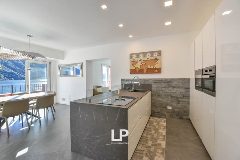 Modern kitchen with central island, marble countertops, granite backsplash, integrated appliances, and a dining area with a view of the balcony and lake.