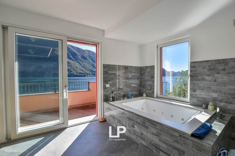 Large walk-in bathtub, two glass doors leading to balcony, scenic views of lake and mountains, tiled walls and floors