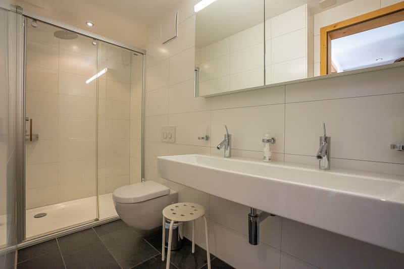 modern bathroom with white tiles, glass shower, white toilet, white sink, white stool, mirror