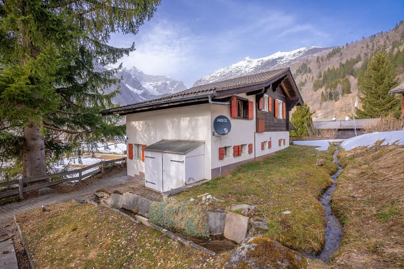 small chalet, white walls, brown roof, red shutters, satellite dish, small garden, snowy mountains in the background