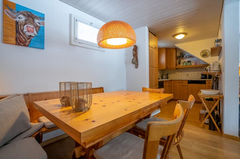 Dining area with wooden table, chairs, and a bench. Wooden cabinets, a sink, and a dishwasher in the kitchen. A painting of a cow on the wall.