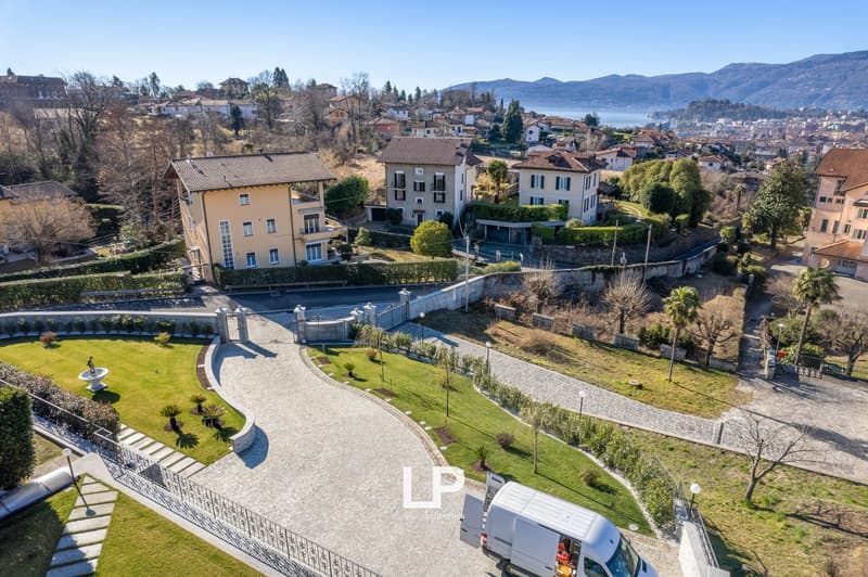 Large driveway leading to house, with several residential buildings in background, green hills, mountains, and water body