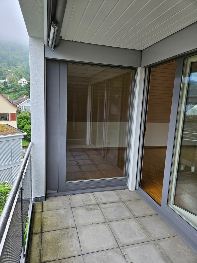 The image shows a balcony or terrace with a sliding glass door leading to the interior. The balcony has a tiled floor and a wooden ceiling with a ventilation system. The surrounding area outside the balcony appears to be a residential neighborhood with tr