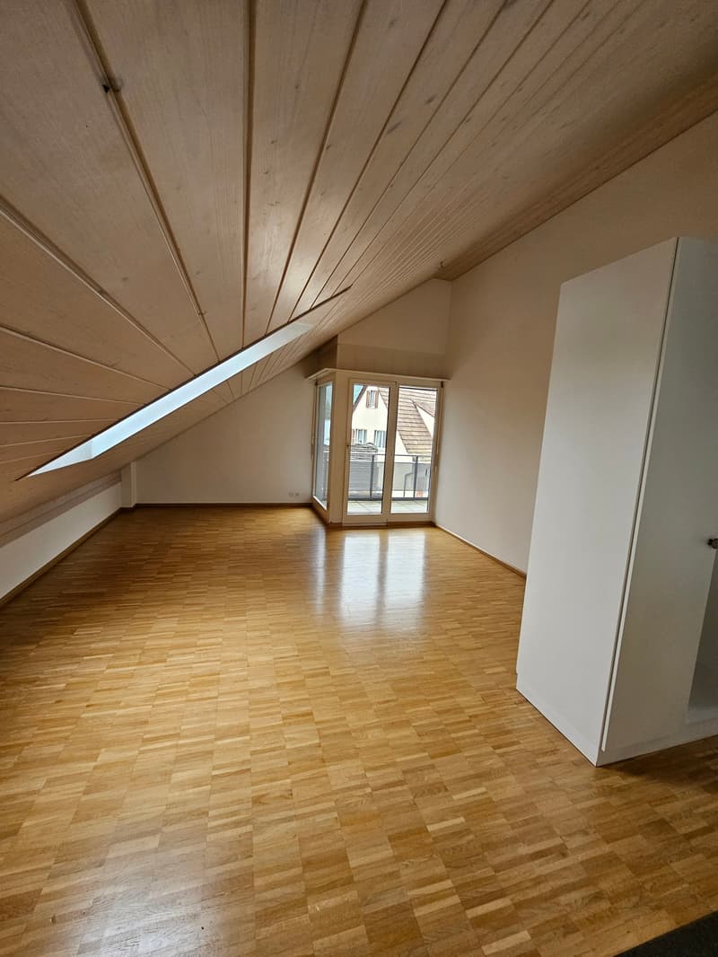 Spacious attic bedroom with wooden ceiling, hardwood flooring, and a balcony/terrace with large windows providing natural light.