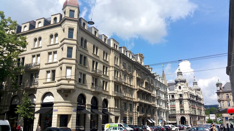 A large, multi-story building with beige stone walls, numerous arched windows, several balconies, and large awnings. The top is rounded with domes. Cars are parked in front of the building.