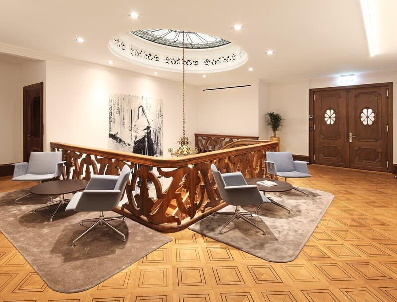 wooden handrail with a circular metal chandelier, wooden double doors, wooden flooring, two wooden chairs, round tables, carpet, white walls, artwork, and a potted plant