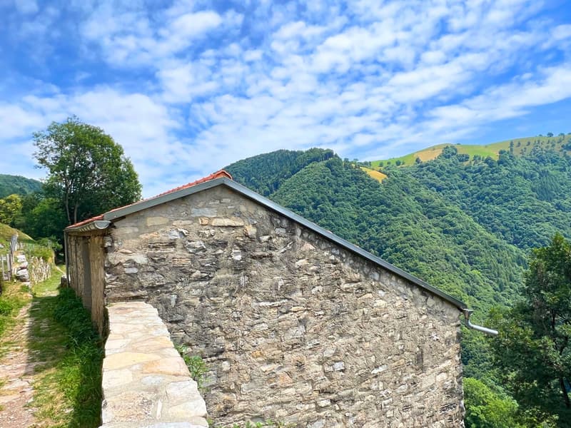 Old stone house with red roof, situated in a mountainous area with lush green vegetation