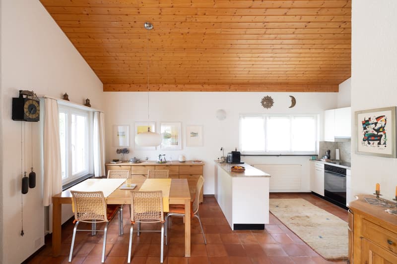 Dining room with wooden table, chairs, kitchen with white cabinets, countertop, and appliances, wooden ceiling, tiled floor, and various decorative items.