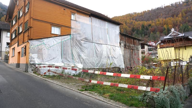 House under construction, wooden exterior, covered with plastic sheeting, surrounded by a fence, road, and greenery.