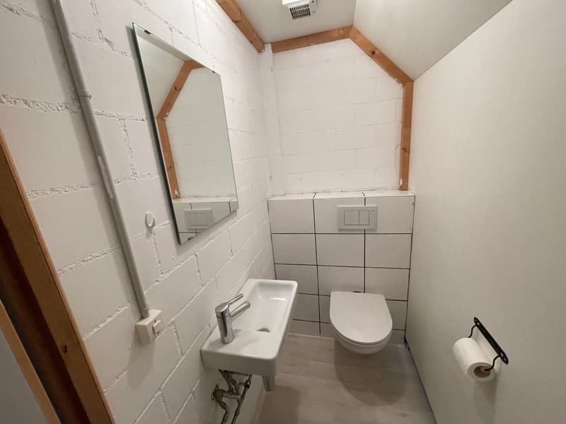 Small bathroom with white tiles, white sink, white toilet, mirror, and wooden ceiling beams.
