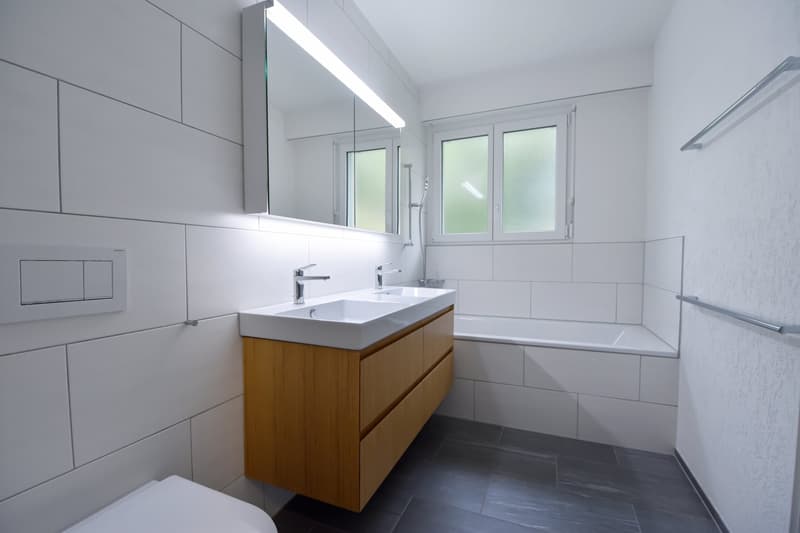 Bathroom with white tiles, wooden vanity, bathtub, modern faucet, shower head, mirror, and towel bars