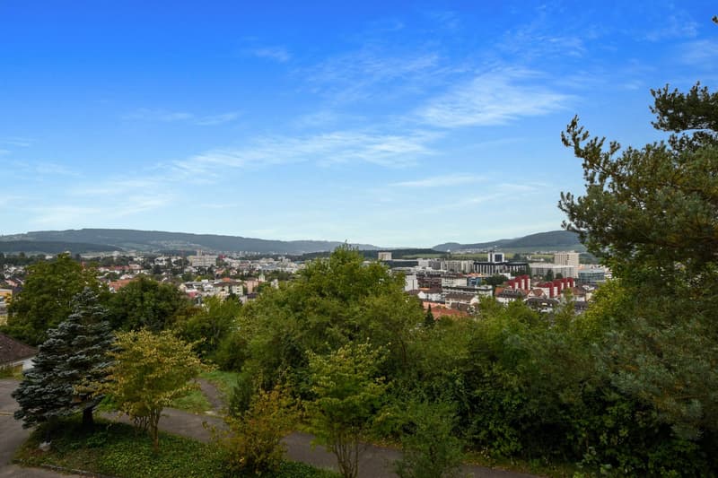 Aerial view of a city surrounded by greenery, with buildings, hills, and a clear blue sky.