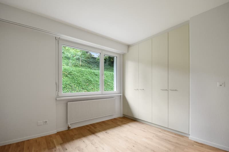 Large window with outside view, white walls, wooden floors, white cabinets, white radiator