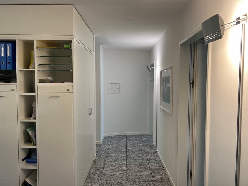 The image shows an office or workspace with a built-in storage cabinet, shelves, and a tiled floor. The room has white walls and appears to be well-lit. There is a door visible at the end of the hallway.