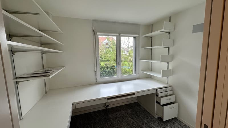 The image shows a bedroom with a large window, white walls, and built-in shelving and storage units along the walls. There is a desk with drawers and a carpeted floor.