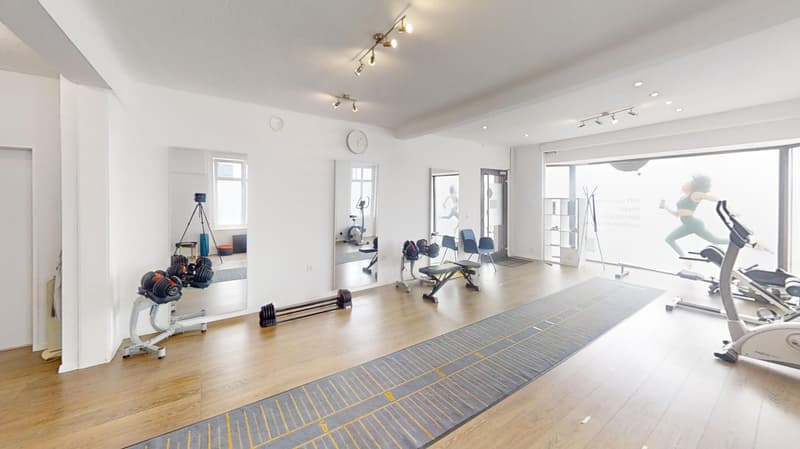 Large open space with hardwood floors, white walls, and various fitness equipment such as dumbbells, weight racks, and exercise machines. The room is well-lit with track lighting and has large windows providing natural light.