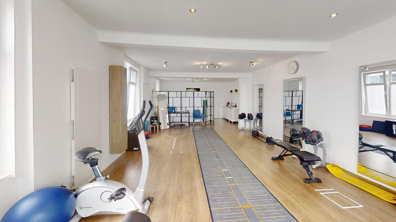 Large open space with hardwood floors, various exercise equipment such as stationary bikes, weight benches, and other fitness gear, shelving units, and a track or marked area on the floor for workouts.
