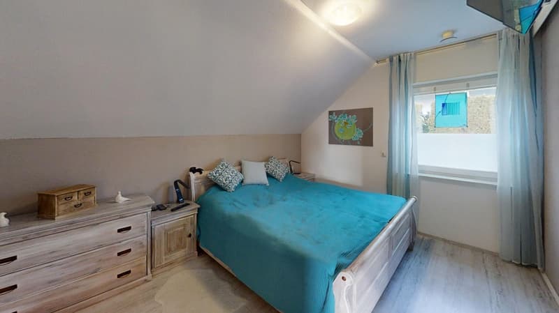 Double bed with blue cover, wooden dressers, nightstands, artwork, window with curtains, slanted ceiling