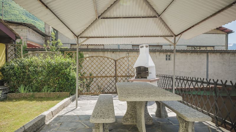 Outdoor dining area with table and benches, covered by a gazebo, stone flooring, garden with plants, wrought iron fence, outdoor fireplace, metal grate