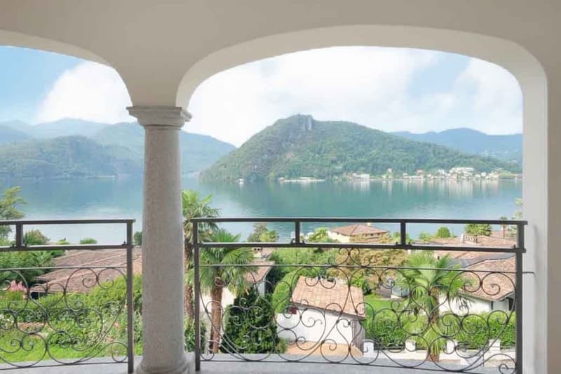 Arched balcony with ornate wrought-iron railing, overlooking a scenic lake surrounded by mountains and lush greenery