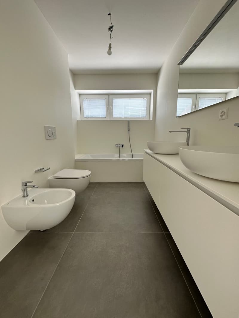 Spacious bathroom with white walls, large windows, tiled floor, bathtub, two sinks, and toilet