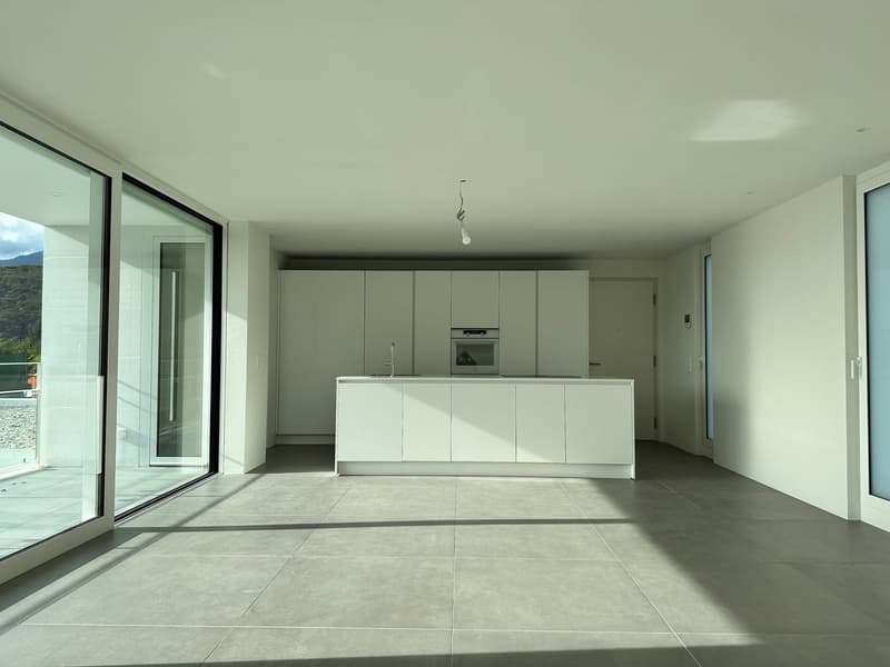 The image shows a spacious, open-concept living area with a modern kitchen. The room features large windows that provide ample natural light and offer a scenic view of the surrounding landscape. The kitchen is equipped with sleek, white cabinets and a cen