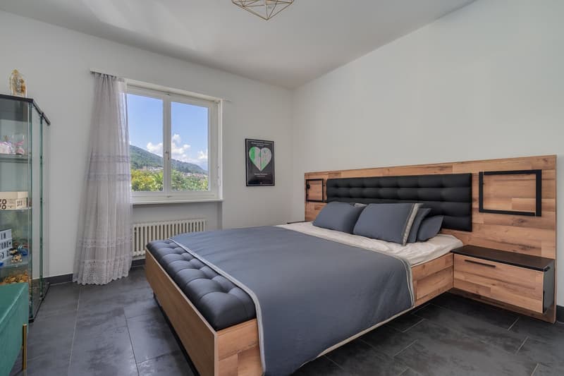 Spacious bedroom with large windows offering a scenic view, wooden bed frame with tufted headboard, gray bedding, and a decorative framed artwork on the wall