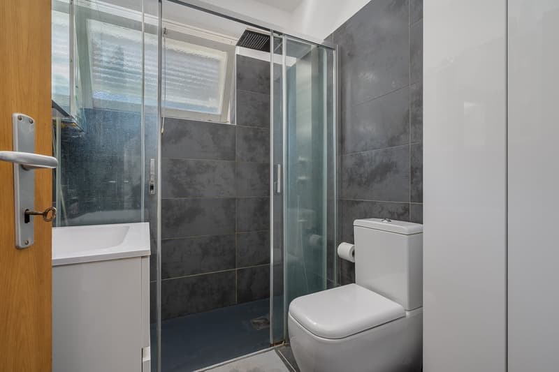 Bathroom with gray tile walls, glass shower enclosure, toilet, and vanity sink