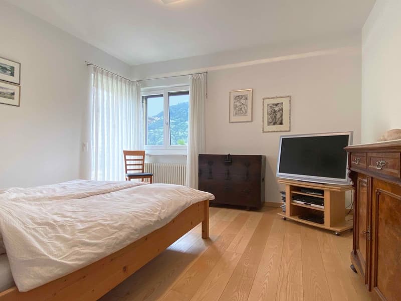 Spacious bedroom with large windows offering a scenic view, hardwood flooring, a wooden bed frame, a TV stand, and framed artwork on the walls.
