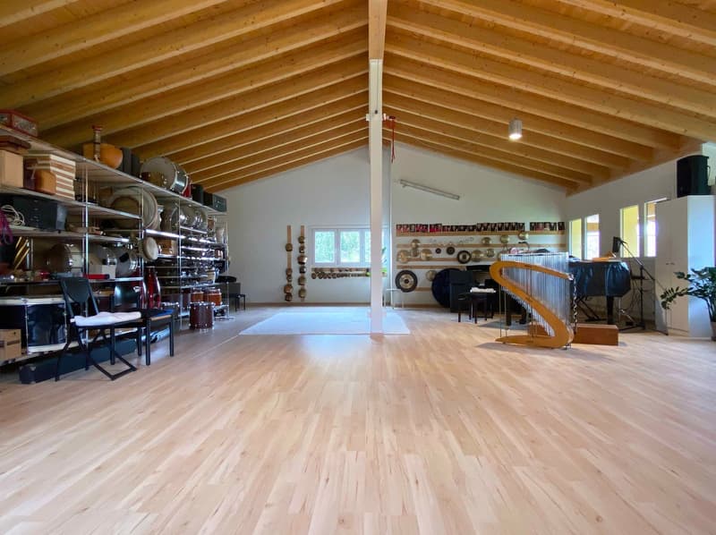 Large open space with wooden beamed ceiling, hardwood floors, shelving units with various items, musical instruments like a piano, and a fireplace visible in the corner.