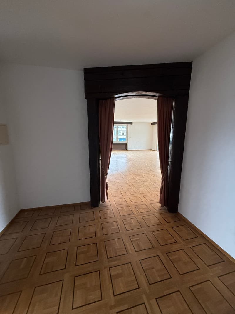 Empty room with wooden parquet flooring, white walls, wooden arch doorway, red curtains