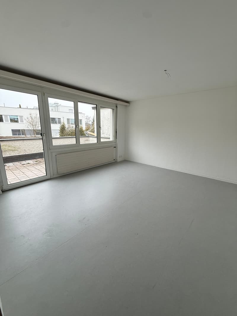 Empty room, white walls, gray floor, large sliding glass doors leading to a balcony, white ceiling, white radiator
