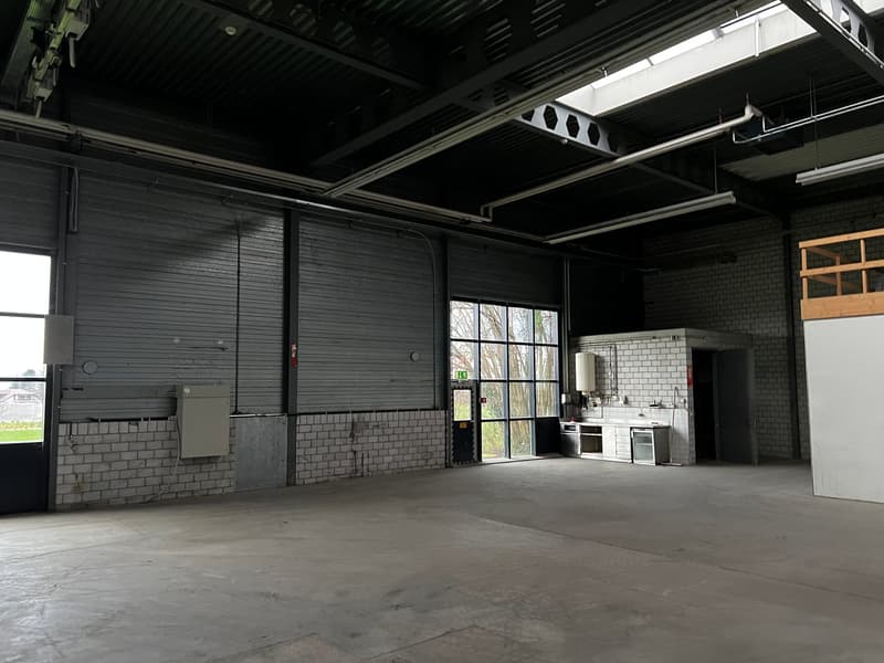 Empty warehouse with brick walls, metal ceiling, large windows, and a small kitchen area.