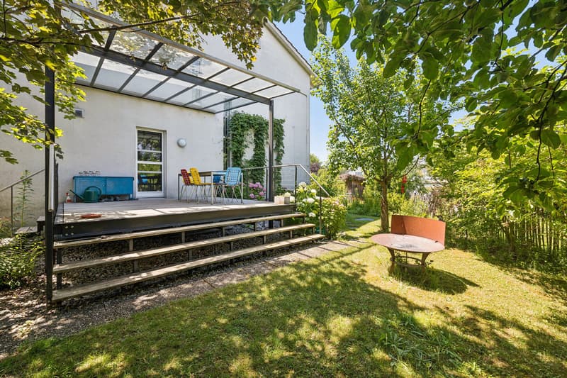 Der Aussenbereich wird durch eine überdachte Terrasse ergänzt, während die abwechslungsreiche Bepflanzung im gesamten Garten für natürliche Beschattung sorgt.