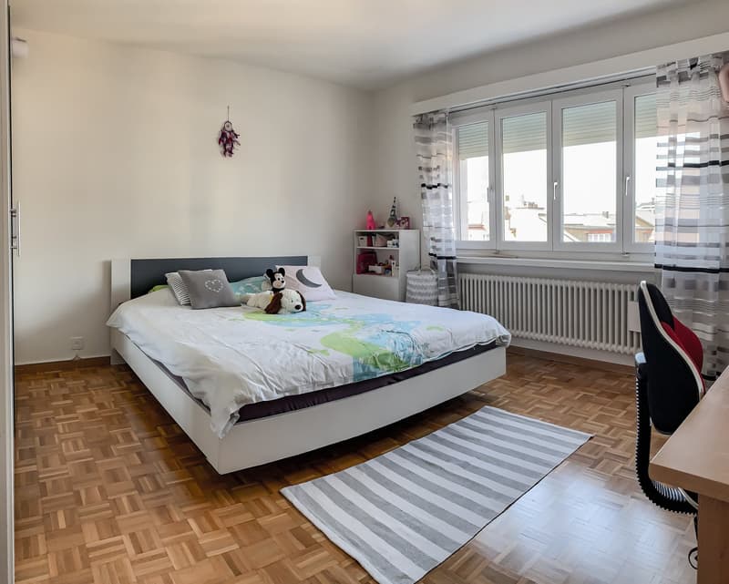 Bedroom with bed, pillows, stuffed toys, rug, white walls, wooden floor, white shelves, radiator, windows with curtains, desk, chair