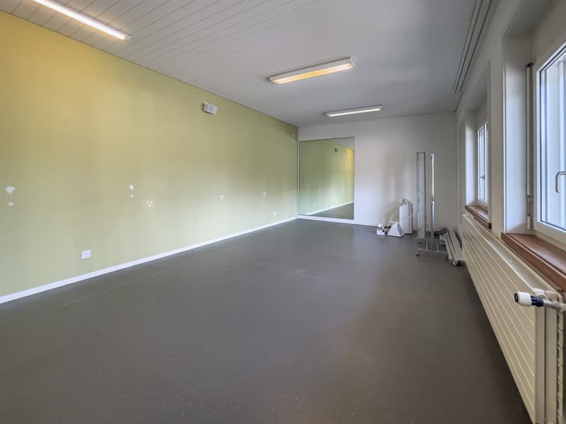 Large empty room with green walls, gray concrete floor, and fluorescent lighting fixtures on the ceiling. There is a window on the side wall and some equipment or furniture items near the window.