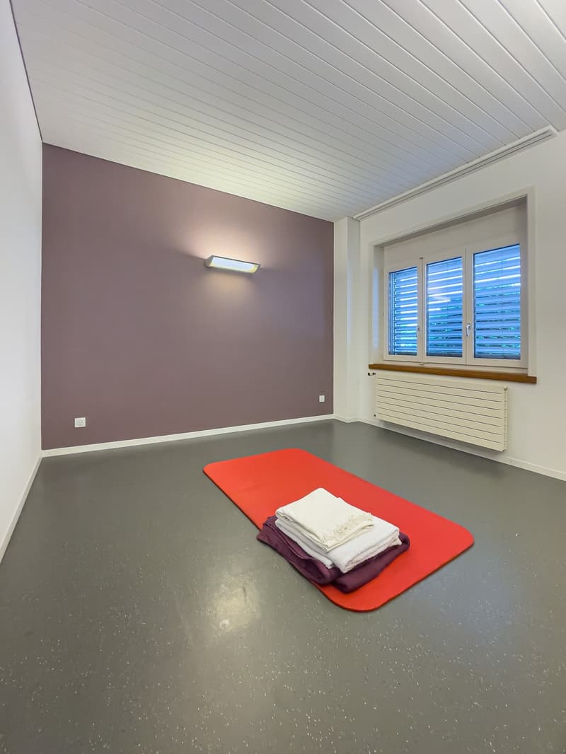This image shows a room with a gray wall, a white ceiling with horizontal panels, and a large window with blinds. There is a red exercise mat on the floor with some folded towels or linens on top. The room appears to be a private space, possibly a home gy