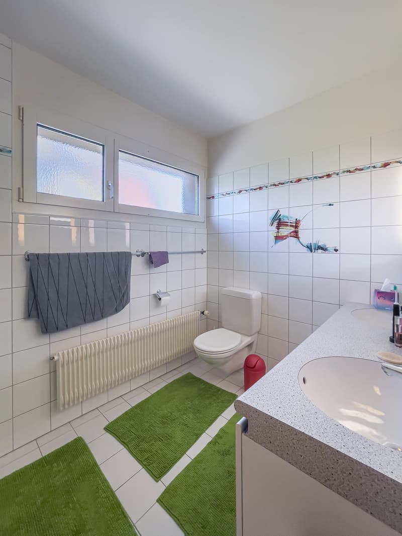 A modern white tiled bathroom with a white sink, a white toilet, and a towel rack with two towels. There is also a white radiator and two green rugs.