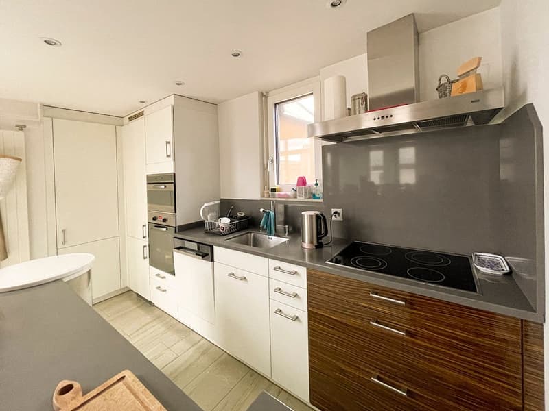 Modern kitchen with white cabinets, wooden drawer fronts, induction hob, extractor hood, stainless steel sink, white countertop, built-in dishwasher, silver appliances, window, and dishwasher